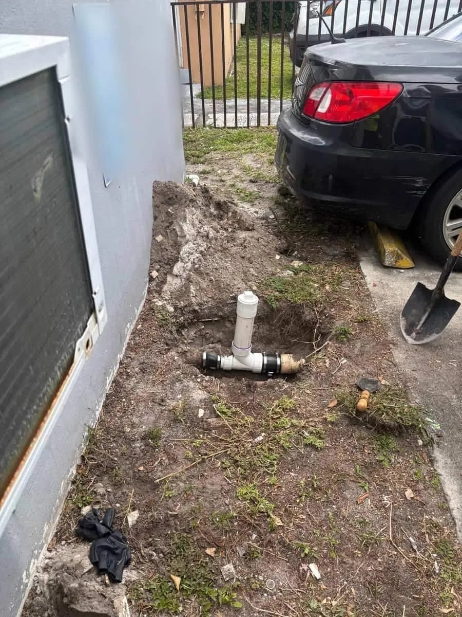 A hand-dug opening along an exterior wall exposing a new white PVC cleanout fitting in the soil