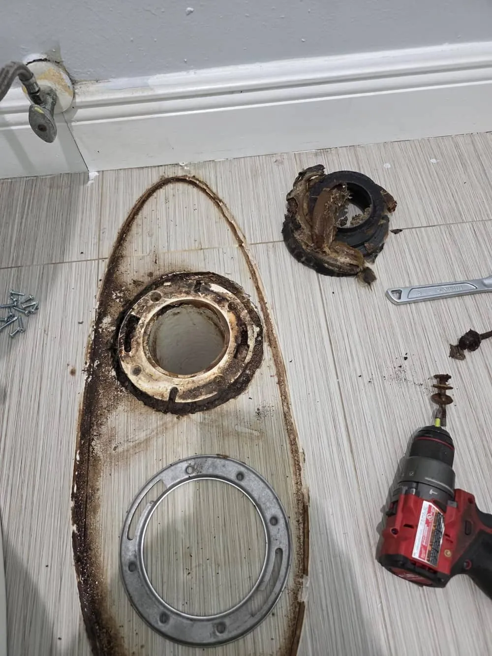 A corroded toilet flange ring removed and lying on the tile floor beside the open drain, with a drill and the old bolts around it