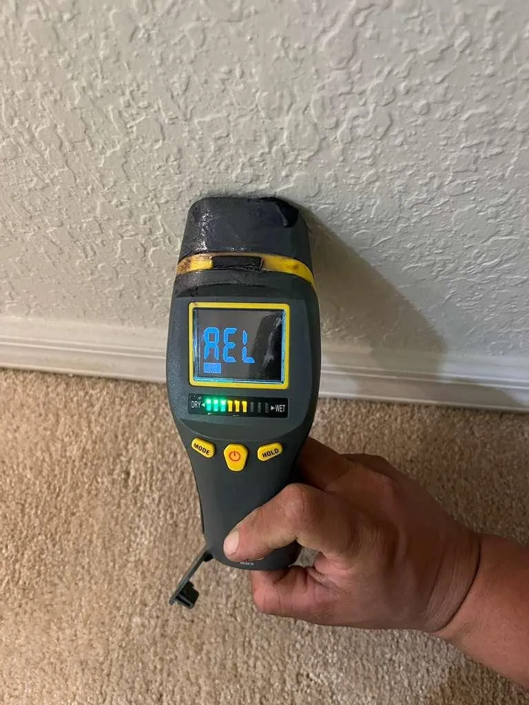 A hand holding a moisture meter with its scale lit while checking a baseboard for hidden water